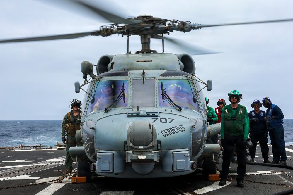 A military helicopter designated '707' and named 'CERBERUS' is on the flight deck of a naval vessel at sea, with its rotors spinning. Several crew members in various colored vests and helmets are positioned around the aircraft, suggesting pre-flight or post-flight operations. The scene takes place on what appears to be a U.S. Navy ship deck with the ocean visible in the background.