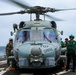 A military helicopter designated '707' and named 'CERBERUS' is on the flight deck of a naval vessel at sea, with its rotors spinning. Several crew members in various colored vests and helmets are positioned around the aircraft, suggesting pre-flight or post-flight operations. The scene takes place on what appears to be a U.S. Navy ship deck with the ocean visible in the background.