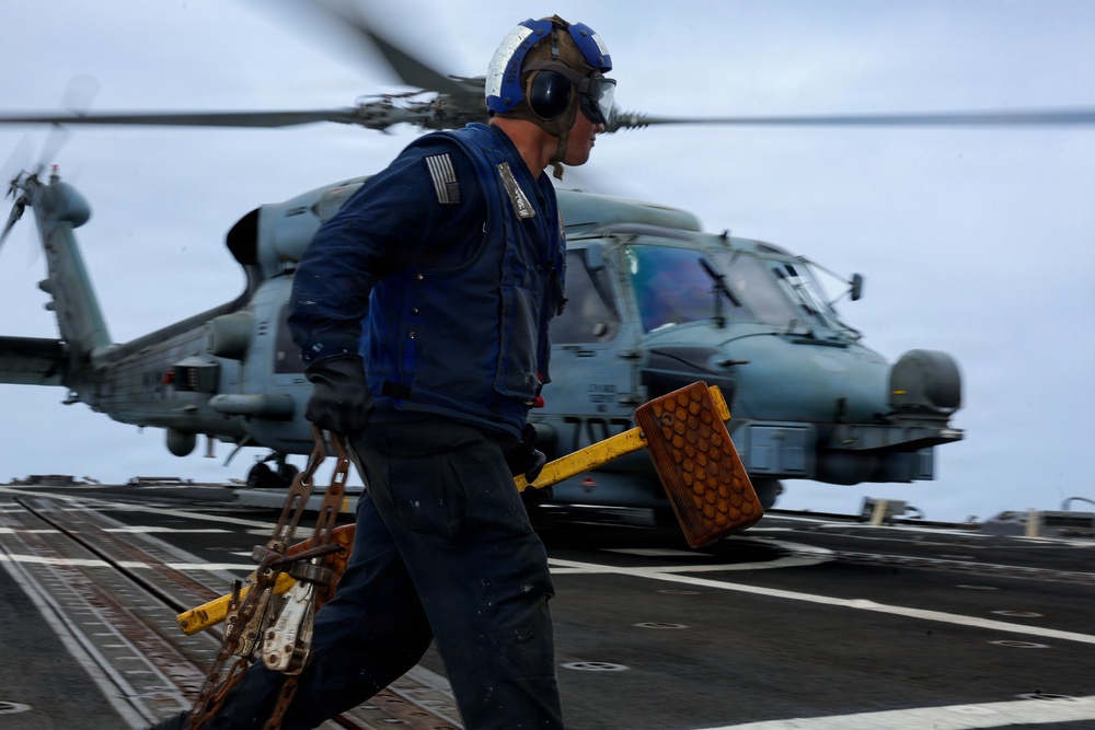 A U.S. Navy deck crew member is working on a ship's flight deck in front of a military helicopter, likely an MH-60 Seahawk. The sailor is carrying wheel chocks and tie-down chains, indicating pre- or post-flight deck operations. The image is taken with motion blur, conveying urgency and dynamic movement in a naval aviation environment.