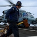 A U.S. Navy deck crew member is working on a ship's flight deck in front of a military helicopter, likely an MH-60 Seahawk. The sailor is carrying wheel chocks and tie-down chains, indicating pre- or post-flight deck operations. The image is taken with motion blur, conveying urgency and dynamic movement in a naval aviation environment.