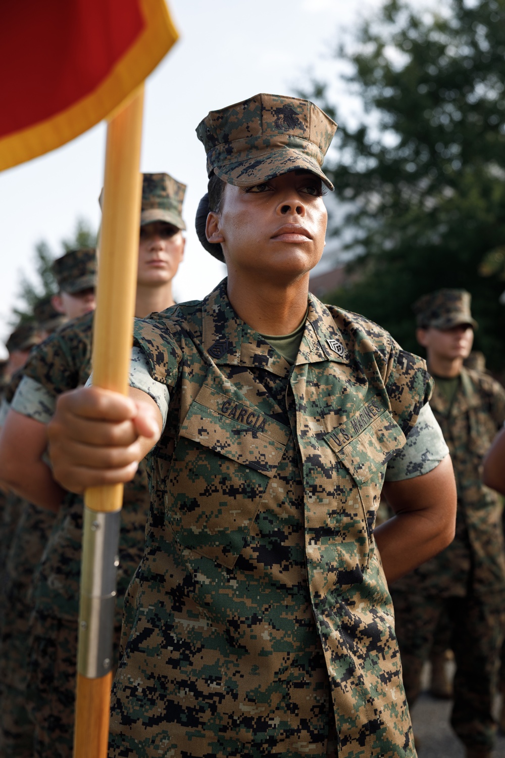 A U.S. Marine identified by the name tape 'Garcia' stands at attention holding a guidon (unit flag) with a red and gold pennant. The Marine is wearing the MARPAT woodland digital camouflage uniform with a cover (cap) bearing the Eagle, Globe, and Anchor emblem. Several other Marines in the same uniform are visible in the background, suggesting a formation or ceremony.