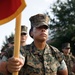 A U.S. Marine identified by the name tape 'Garcia' stands at attention holding a guidon (unit flag) with a red and gold pennant. The Marine is wearing the MARPAT woodland digital camouflage uniform with a cover (cap) bearing the Eagle, Globe, and Anchor emblem. Several other Marines in the same uniform are visible in the background, suggesting a formation or ceremony.