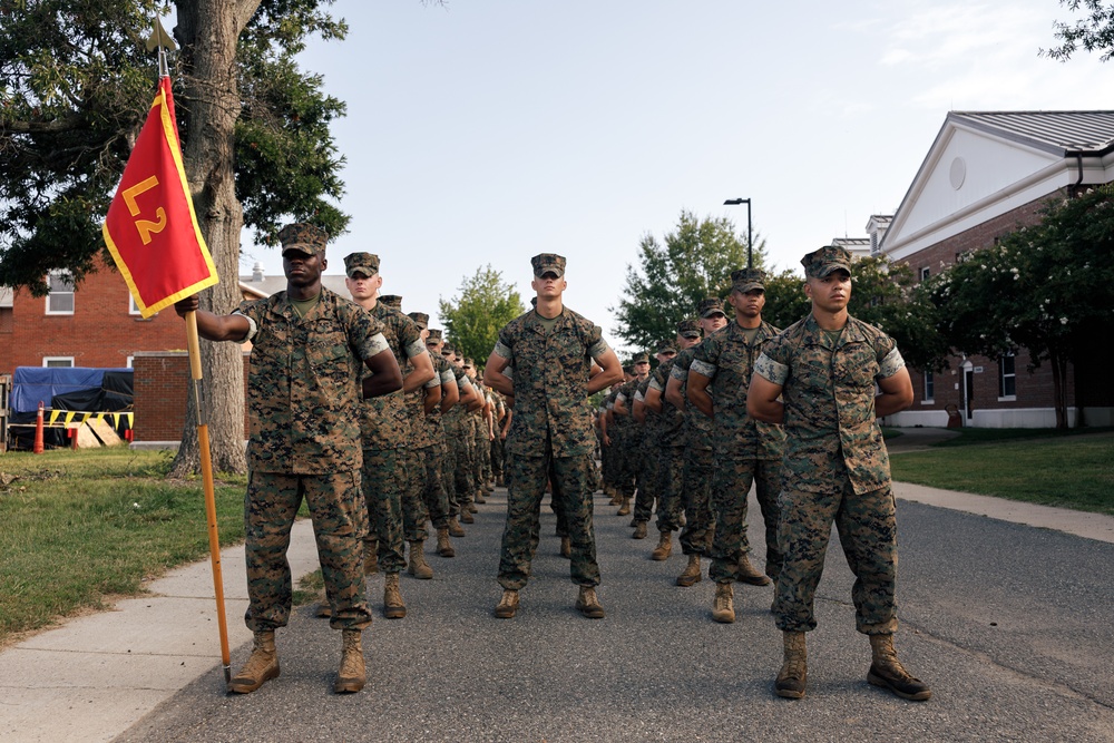 A formation of U.S. Marines in woodland MARPAT camouflage uniforms stands at parade rest on a paved road at what appears to be a military installation. The Marines are arranged in multiple columns, with the leftmost Marine holding a red guidon flag marked 'L2', indicating Lima Company, 2nd Battalion or similar unit designation. The setting includes brick buildings and mature trees consistent with an established military base.