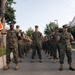 A formation of U.S. Marines in woodland MARPAT camouflage uniforms stands at parade rest on a paved road at what appears to be a military installation. The Marines are arranged in multiple columns, with the leftmost Marine holding a red guidon flag marked 'L2', indicating Lima Company, 2nd Battalion or similar unit designation. The setting includes brick buildings and mature trees consistent with an established military base.
