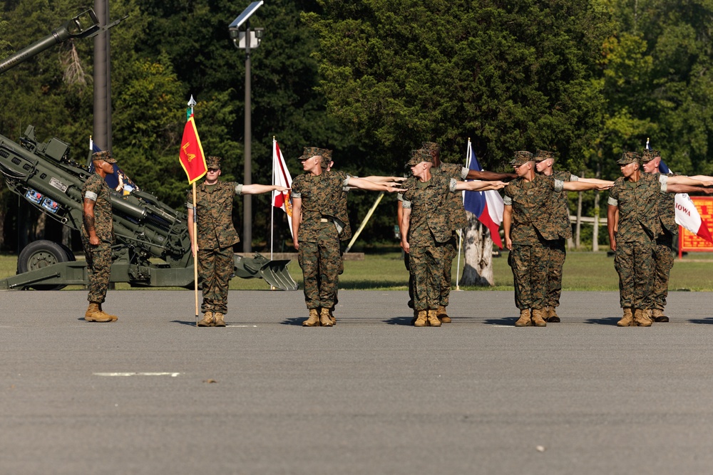 A group of U.S. Marines in woodland MARPAT camouflage uniforms are performing a dress alignment drill on a paved parade ground. Personnel are extending their arms to maintain proper spacing in a formal military formation. A howitzer artillery piece is visible on the left side, and multiple guidon flags including ones labeled 'LA' and 'IOWA' are present.