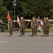 A group of U.S. Marines in woodland MARPAT camouflage uniforms are performing a dress alignment drill on a paved parade ground. Personnel are extending their arms to maintain proper spacing in a formal military formation. A howitzer artillery piece is visible on the left side, and multiple guidon flags including ones labeled 'LA' and 'IOWA' are present.