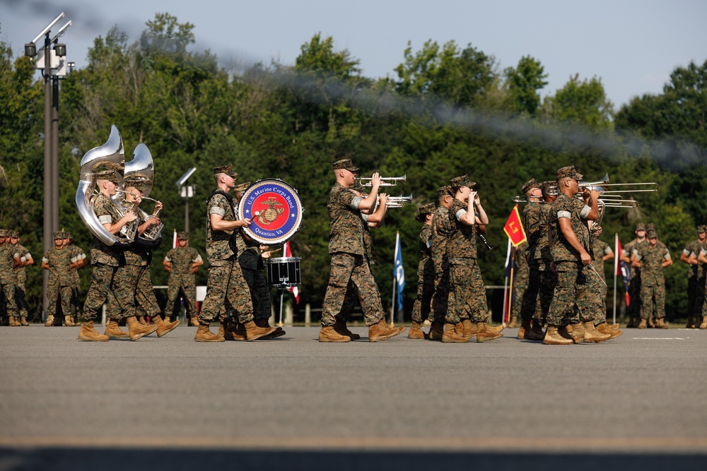 The image shows the U.S. Marine Corps Band from Quantico, VA performing during an outdoor military parade or ceremony. Band members dressed in woodland MARPAT camouflage uniforms are marching in formation while playing brass and percussion instruments. Additional Marines in formation are visible in the background, along with military flags and a smoke trail in the sky.