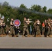 The image shows the U.S. Marine Corps Band from Quantico, VA performing during an outdoor military parade or ceremony. Band members dressed in woodland MARPAT camouflage uniforms are marching in formation while playing brass and percussion instruments. Additional Marines in formation are visible in the background, along with military flags and a smoke trail in the sky.