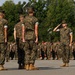 A large formation of military personnel in MARPAT (Marine pattern) camouflage uniforms are assembled on a parade deck or drill field. One service member in the foreground is rendering a salute, while others stand at attention facing away from the camera. The scene appears to be a formal military ceremony or formation event, likely at a U.S. Marine Corps installation.