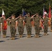 A group of U.S. Marines in woodland MARPAT camouflage uniforms are standing at attention and rendering a salute during a formal military ceremony on a paved parade deck. Multiple flags are visible including the U.S. flag, Marine Corps colors, and unit guidons. Military equipment, including what appears to be an artillery piece or vehicle, is visible in the background.