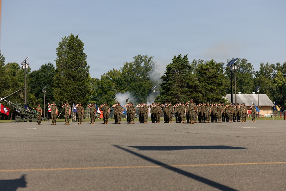 A military formation of personnel in camouflage uniforms stands at salute on a paved parade ground during a formal ceremony. Smoke is visible in the background, consistent with a cannon or artillery salute firing. Multiple unit flags and guidons are present on the left side of the formation.