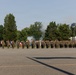 A military formation of personnel in camouflage uniforms stands at salute on a paved parade ground during a formal ceremony. Smoke is visible in the background, consistent with a cannon or artillery salute firing. Multiple unit flags and guidons are present on the left side of the formation.