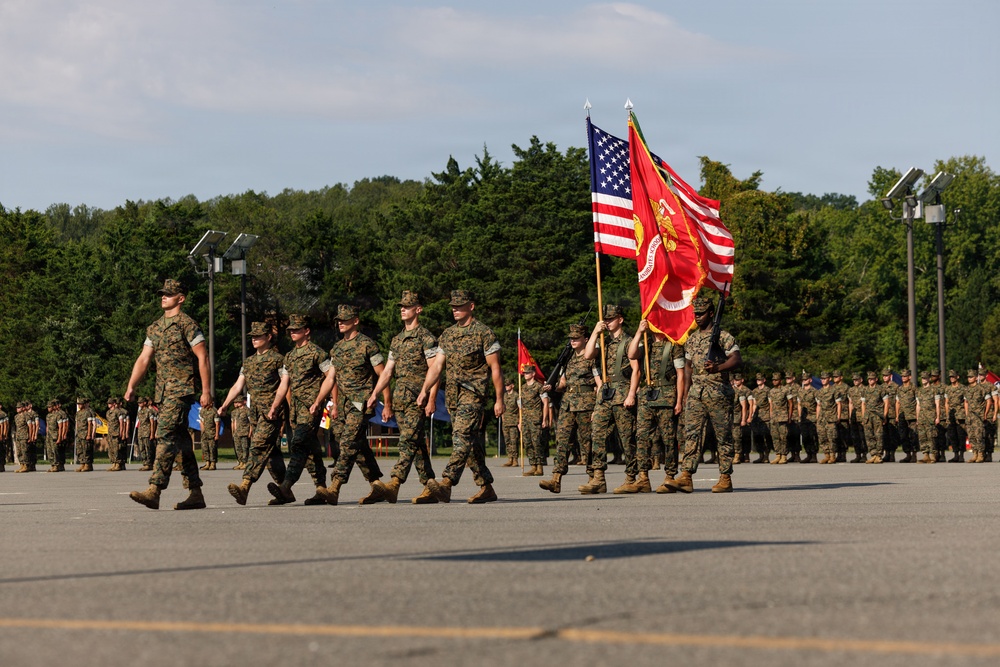A U.S. Marine Corps military parade or ceremony is taking place on a large paved parade deck. A color guard is prominently carrying the American flag and the Marine Corps flag, while formations of Marines in woodland MARPAT camouflage uniforms stand at attention in the background. The setting appears to be a military installation surrounded by trees under partly cloudy skies.