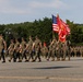 A U.S. Marine Corps military parade or ceremony is taking place on a large paved parade deck. A color guard is prominently carrying the American flag and the Marine Corps flag, while formations of Marines in woodland MARPAT camouflage uniforms stand at attention in the background. The setting appears to be a military installation surrounded by trees under partly cloudy skies.