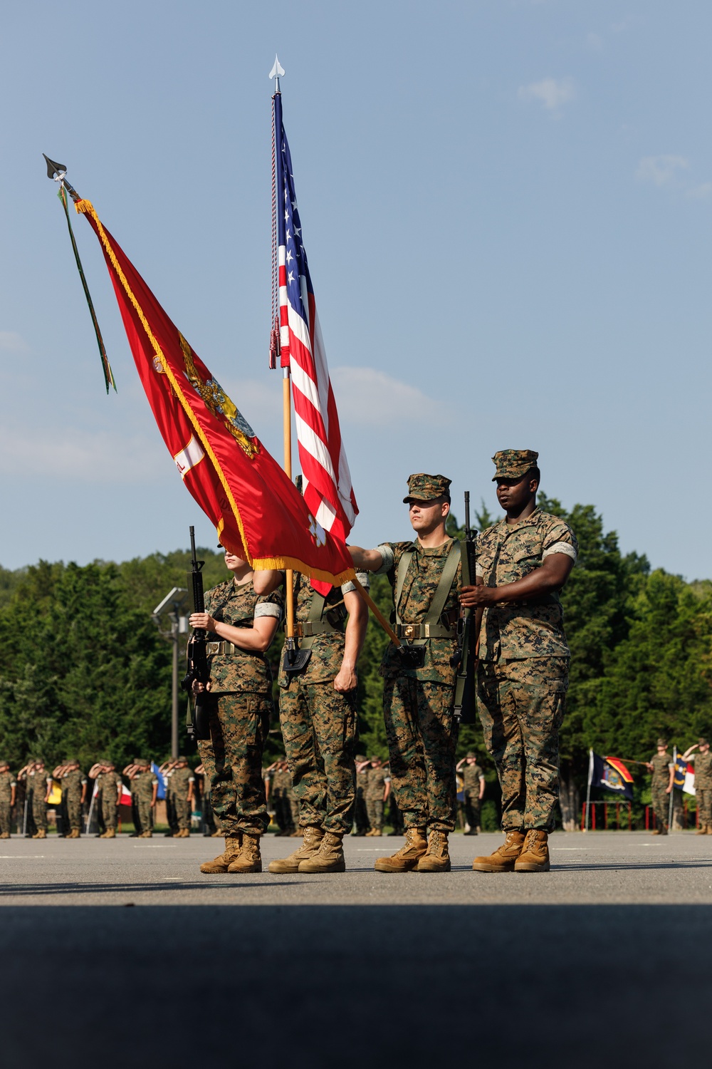 A U.S. Marine Corps color guard stands at attention during a formal military ceremony on a parade deck. Four Marines in MARPAT digital camouflage uniforms carry the U.S. national flag and Marine Corps unit colors, while armed escorts hold rifles. A large formation of Marines and spectators is visible in the background, with many saluting.