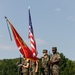 A U.S. Marine Corps color guard stands at attention during a formal military ceremony on a parade deck. Four Marines in MARPAT digital camouflage uniforms carry the U.S. national flag and Marine Corps unit colors, while armed escorts hold rifles. A large formation of Marines and spectators is visible in the background, with many saluting.