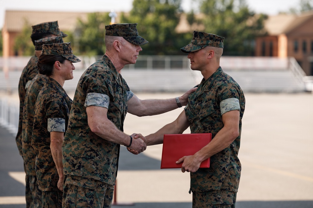 A military ceremony is taking place outdoors on a paved surface, likely at a U.S. Marine Corps installation. A senior Marine officer is shaking hands with a younger Marine while placing a hand on his shoulder, suggesting a formal recognition or award presentation. The recipient is holding a red folder, commonly used to present certificates or commendations.