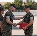 A military ceremony is taking place outdoors on a paved surface, likely at a U.S. Marine Corps installation. A senior Marine officer is shaking hands with a younger Marine while placing a hand on his shoulder, suggesting a formal recognition or award presentation. The recipient is holding a red folder, commonly used to present certificates or commendations.