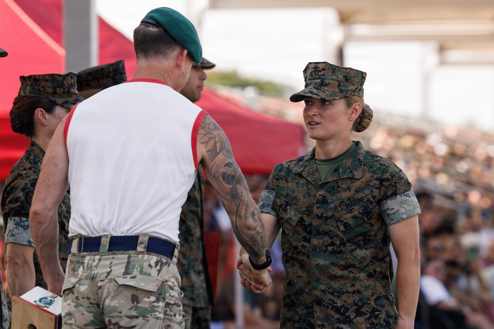 A U.S. Marine in MARPAT digital camouflage uniform is shaking hands with a military personnel wearing a green beret and multicam trousers with a white athletic shirt, suggesting an awards or recognition ceremony. The event appears to be a large public military gathering with a significant crowd visible in the background. Multiple service members from different branches or nations are present, indicating a joint or multinational military event.