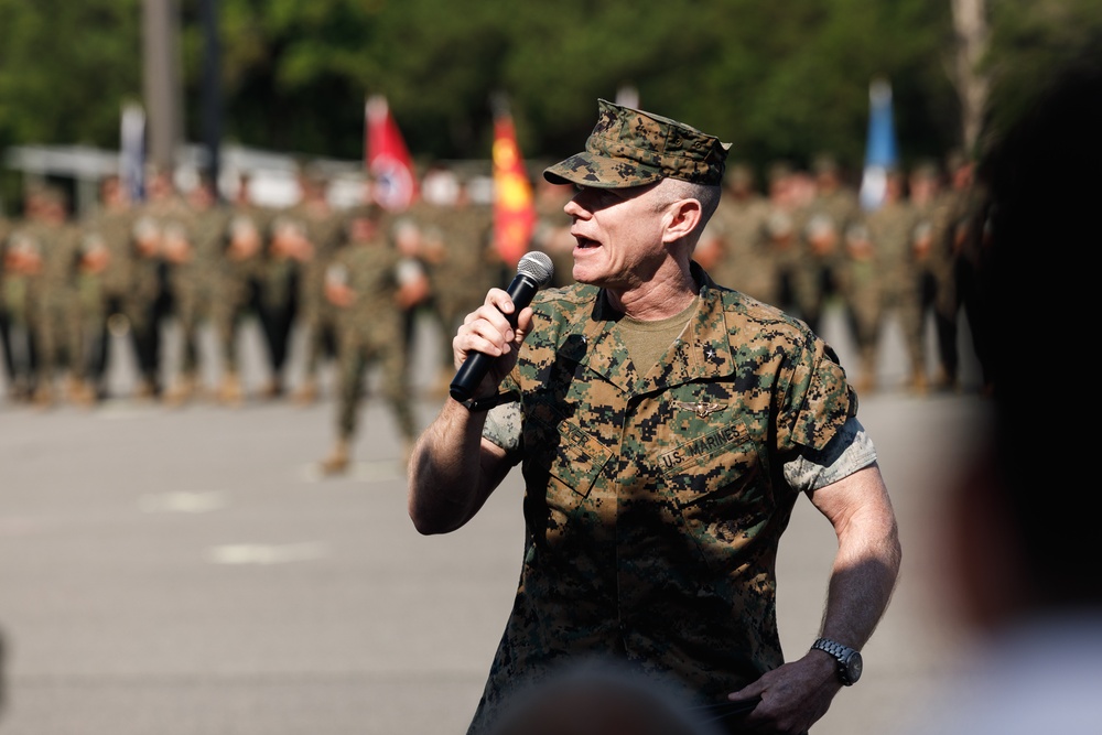 A U.S. Marines officer in MARPAT woodland camouflage uniform is speaking into a handheld microphone, apparently addressing a formation of troops. The speaker appears to be a senior officer, visible by insignia on the uniform, standing outdoors on a paved surface. In the background, multiple Marines stand in formation alongside what appear to be unit guidons or flags.