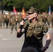 A U.S. Marines officer in MARPAT woodland camouflage uniform is speaking into a handheld microphone, apparently addressing a formation of troops. The speaker appears to be a senior officer, visible by insignia on the uniform, standing outdoors on a paved surface. In the background, multiple Marines stand in formation alongside what appear to be unit guidons or flags.