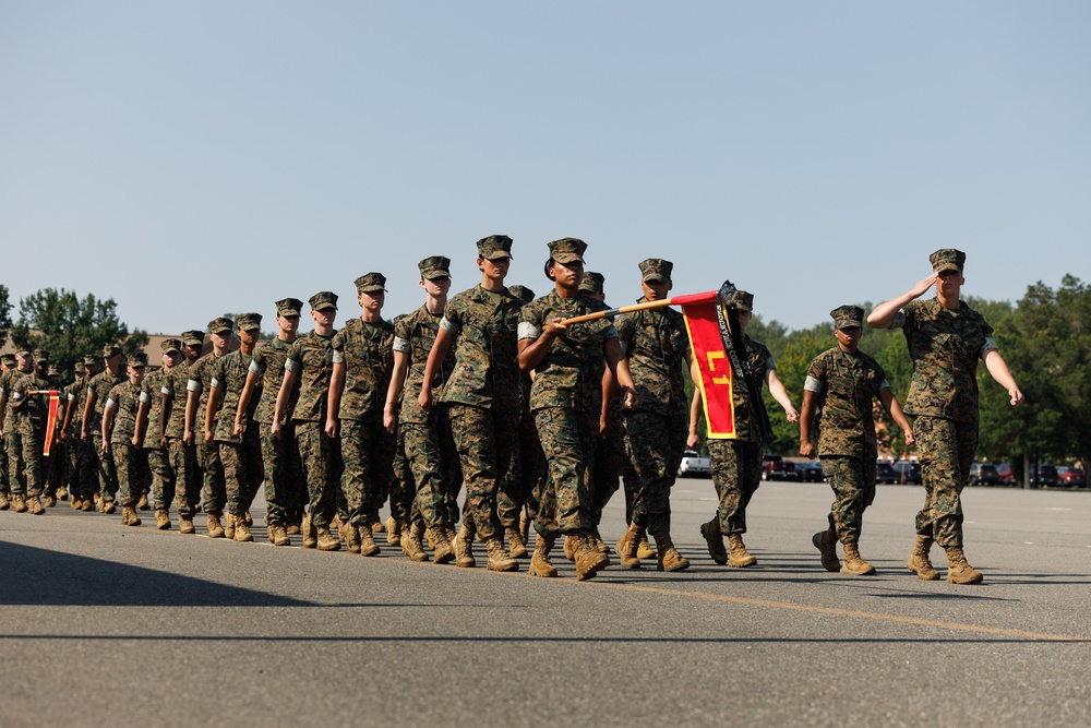 A formation of U.S. Marines in digital woodland MARPAT camouflage uniforms marches in a parade or graduation ceremony on a paved surface. A guidon bearer carries a red and gold unit pennant near the front of the formation. The scene appears to take place at a military installation, likely a recruit training depot or base parade deck.