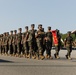 A formation of U.S. Marines in digital woodland MARPAT camouflage uniforms marches in a parade or graduation ceremony on a paved surface. A guidon bearer carries a red and gold unit pennant near the front of the formation. The scene appears to take place at a military installation, likely a recruit training depot or base parade deck.