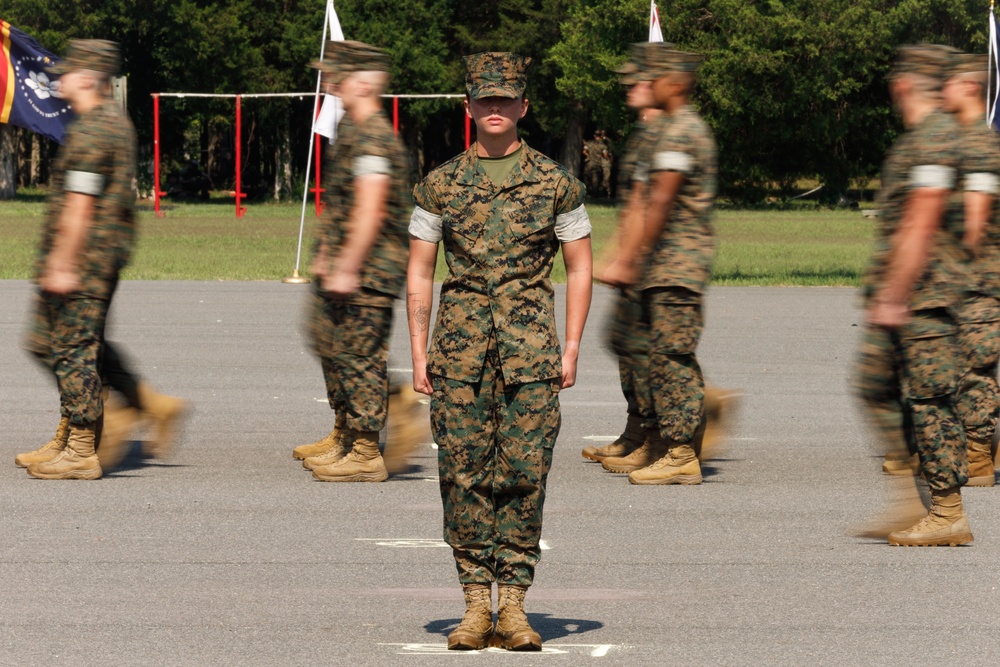 A military formation drill or parade is taking place on a paved surface, likely at a U.S. Marine Corps base. One central figure stands sharply in focus at attention while surrounding personnel are motion-blurred, suggesting they are marching past. The uniforms and digital camouflage pattern are consistent with USMC MARPAT (Marine Pattern) combat utility uniforms.