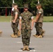 A military formation drill or parade is taking place on a paved surface, likely at a U.S. Marine Corps base. One central figure stands sharply in focus at attention while surrounding personnel are motion-blurred, suggesting they are marching past. The uniforms and digital camouflage pattern are consistent with USMC MARPAT (Marine Pattern) combat utility uniforms.