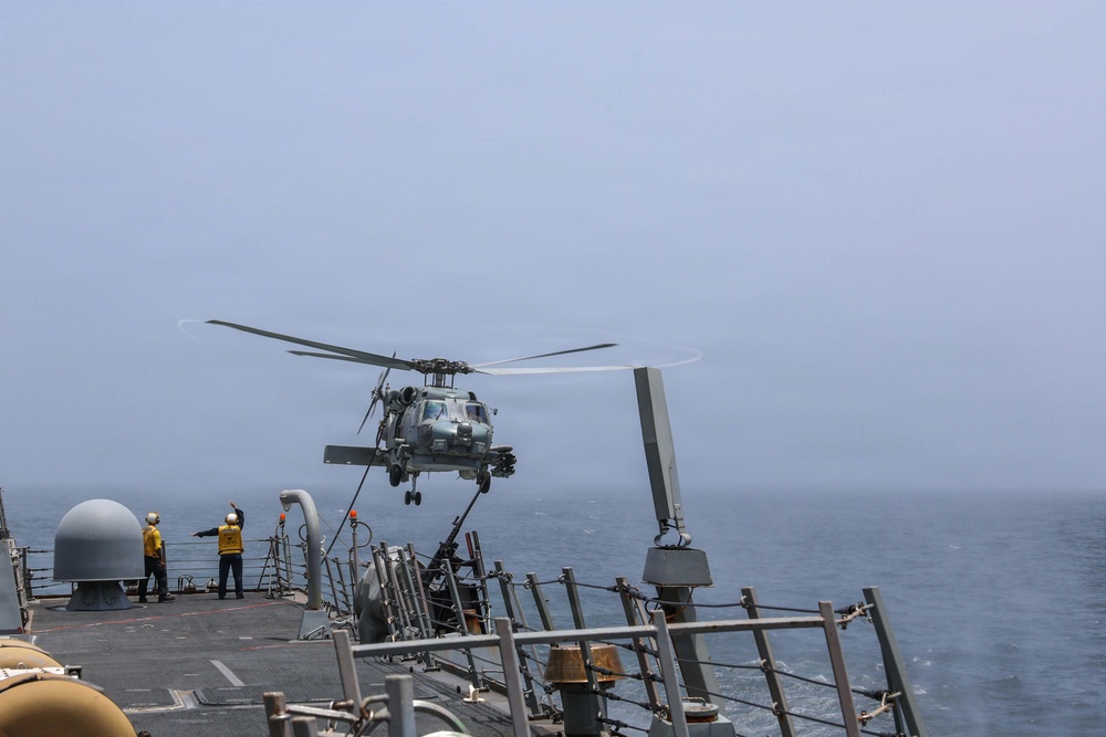 A military helicopter, consistent with an MH-60 Seahawk, is hovering or landing on the flight deck of a naval warship, likely a U.S. Navy destroyer or cruiser. Deck crew members in yellow and white jerseys are visible on the flight deck, with one appearing to signal the helicopter. The scene takes place at sea under overcast, hazy conditions.