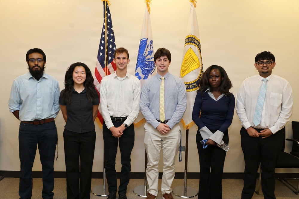 Six young adults are posed together in a formal group photo in front of three flags, including the United States flag and what appears to be a U.S. Department of Commerce or similar federal agency flag. The individuals are dressed in business or business-casual attire, suggesting an official or professional occasion such as an internship, award ceremony, or government program. The setting appears to be an indoor government or institutional facility.
