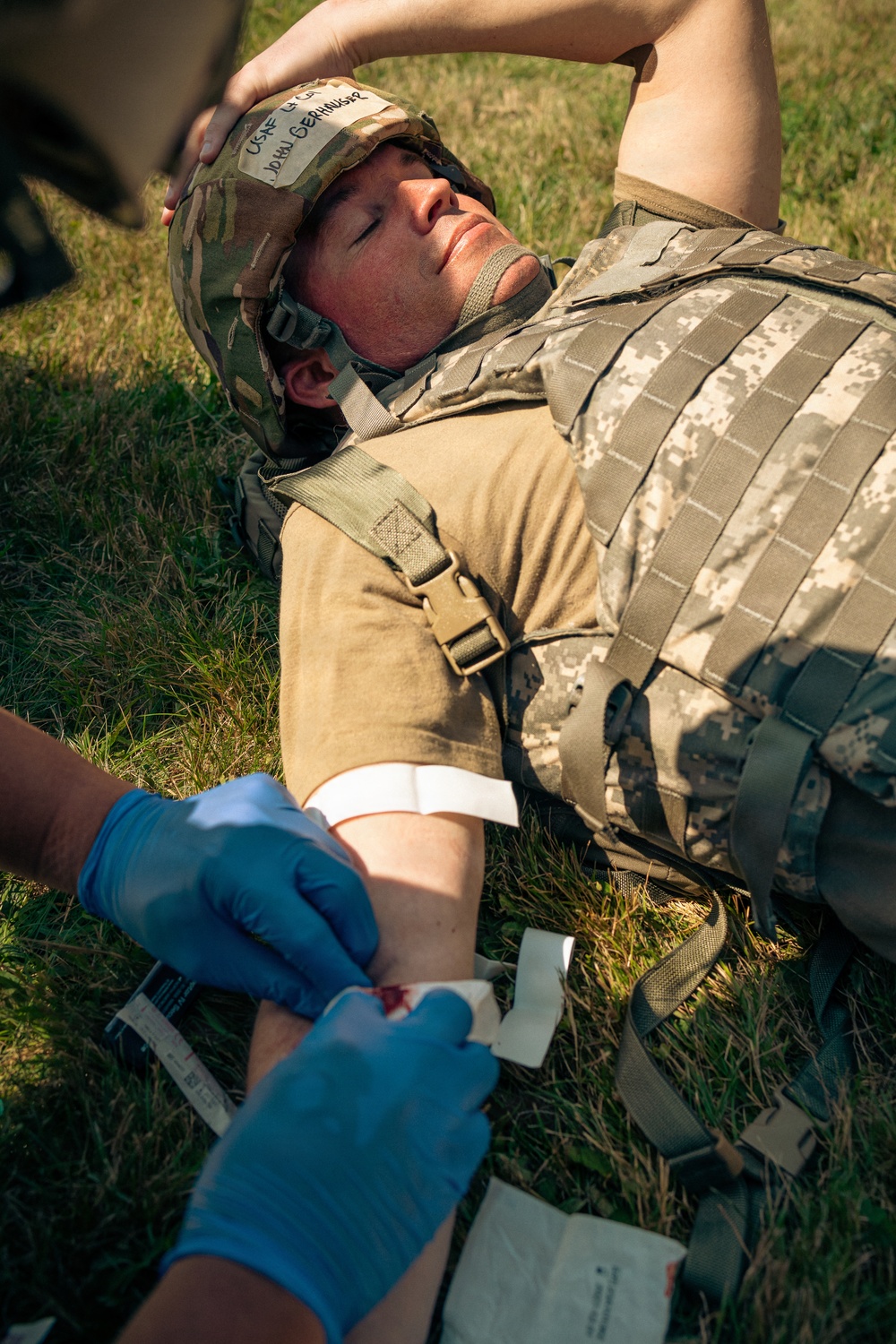 A military personnel is lying on grass receiving field medical treatment on their arm, with a gloved medic applying bandaging or wound care. The subject is wearing a camouflage helmet with a name tag reading 'USAF' and 'John Gerhauser' written in marker, along with a tactical plate carrier vest. This appears to be a combat casualty care training exercise or real field medical scenario.