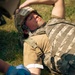 A military personnel is lying on grass receiving field medical treatment on their arm, with a gloved medic applying bandaging or wound care. The subject is wearing a camouflage helmet with a name tag reading 'USAF' and 'John Gerhauser' written in marker, along with a tactical plate carrier vest. This appears to be a combat casualty care training exercise or real field medical scenario.
