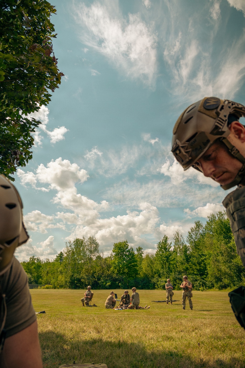 Military personnel in tactical gear are conducting what appears to be a field training exercise on an open grassy area. In the foreground, a soldier wearing a tactical helmet and body armor is visible in close-up, while in the background several soldiers are gathered in small groups, with some kneeling around what may be a simulated casualty scenario. The setting is a sunny day with scattered clouds over a green field bordered by trees.
