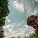 Military personnel in tactical gear are conducting what appears to be a field training exercise on an open grassy area. In the foreground, a soldier wearing a tactical helmet and body armor is visible in close-up, while in the background several soldiers are gathered in small groups, with some kneeling around what may be a simulated casualty scenario. The setting is a sunny day with scattered clouds over a green field bordered by trees.
