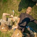 Two military personnel in OCP (Operational Camouflage Pattern) uniforms and tan helmets are conducting tactical combat casualty care (TCCC) training on a medical simulation mannequin. The mannequin, branded 'Operative Experience,' is lying on grass while trainees practice applying a tourniquet and other medical interventions. This appears to be a field medical training exercise.