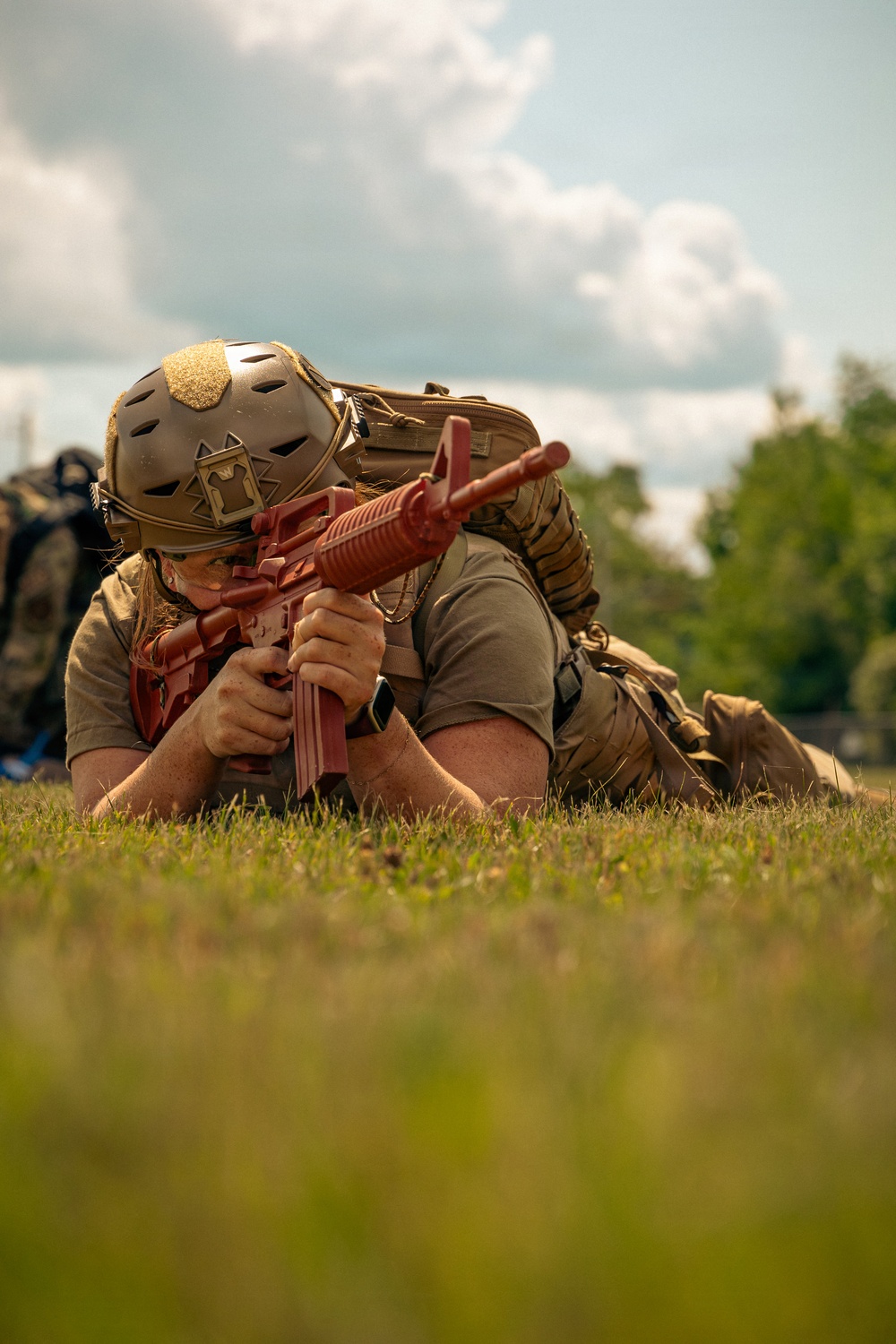 A person in tactical gear is lying prone on grass, aiming a red training/dummy rifle. They are wearing a tan tactical helmet with ventilation holes and a hook-and-loop patch, a plate carrier vest, and carrying a large rucksack. The red-colored firearm indicates this is a training replica or inert dummy weapon used for drills.