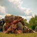 A person in tactical gear is lying prone on grass, aiming a red training/dummy rifle. They are wearing a tan tactical helmet with ventilation holes and a hook-and-loop patch, a plate carrier vest, and carrying a large rucksack. The red-colored firearm indicates this is a training replica or inert dummy weapon used for drills.
