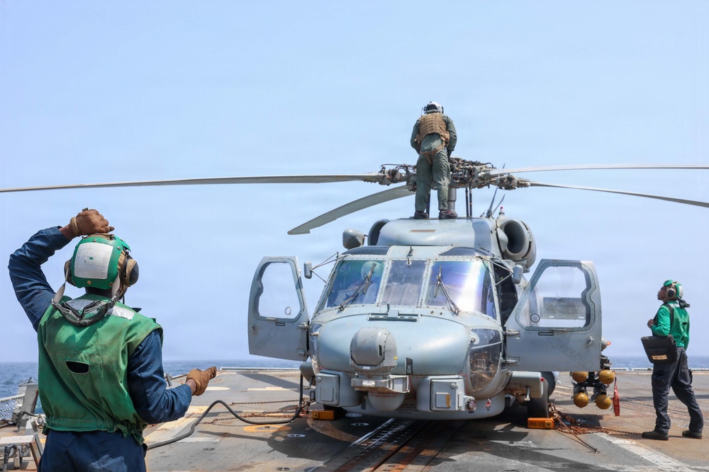 A military helicopter, consistent with an MH-60 Seahawk variant, is positioned on the flight deck of a naval vessel at sea. Deck crew members wearing green jerseys and cranial helmets are conducting maintenance or pre-flight operations around the aircraft. One person is standing on top of the helicopter near the rotor assembly, while others signal or observe from the deck.