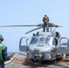 A military helicopter, consistent with an MH-60 Seahawk variant, is positioned on the flight deck of a naval vessel at sea. Deck crew members wearing green jerseys and cranial helmets are conducting maintenance or pre-flight operations around the aircraft. One person is standing on top of the helicopter near the rotor assembly, while others signal or observe from the deck.