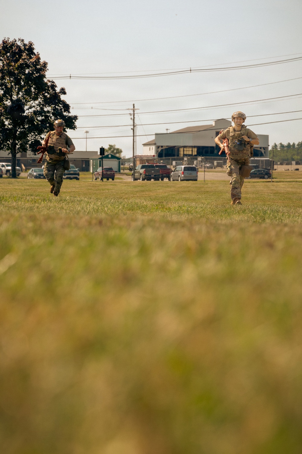 Two military personnel in full combat gear, including helmets and tactical vests, are running across an open grassy field. Both are carrying what appear to be training weapons with orange/red markings, indicating a military exercise or training scenario. The background shows a military installation with buildings, vehicles, and utility lines.