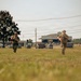 Two military personnel in full combat gear, including helmets and tactical vests, are running across an open grassy field. Both are carrying what appear to be training weapons with orange/red markings, indicating a military exercise or training scenario. The background shows a military installation with buildings, vehicles, and utility lines.