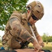 A military or tactical personnel wearing full combat gear is performing a medical training exercise on a mannequin or training dummy in an outdoor field setting. The individual is applying what appears to be a bandage or wound packing to the torso area of the training casualty. This appears to be a Tactical Combat Casualty Care (TCCC) or similar battlefield medical training scenario.