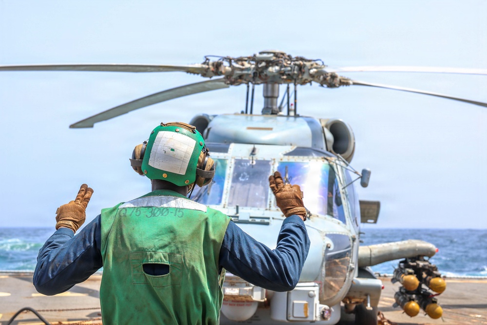 A naval deck crew member wearing a green safety vest and helmet with ear protection is signaling with both hands raised toward a military helicopter on what appears to be a ship's flight deck. The helicopter, painted in a light gray naval color scheme, has its rotor blades extended and appears to be preparing for takeoff or has just landed. The scene takes place at sea, with open ocean visible in the background.