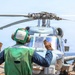 A naval deck crew member wearing a green safety vest and helmet with ear protection is signaling with both hands raised toward a military helicopter on what appears to be a ship's flight deck. The helicopter, painted in a light gray naval color scheme, has its rotor blades extended and appears to be preparing for takeoff or has just landed. The scene takes place at sea, with open ocean visible in the background.
