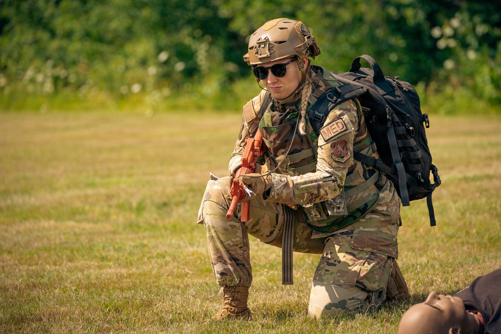A female military medic in full combat gear kneels on a grass field, appearing to attend to a casualty partially visible at the bottom right of the frame. She is wearing OCP (Operational Camouflage Pattern) uniform with a 'MED' patch visible on her shoulder, a tan tactical helmet, sunglasses, and carries a black MOLLE medical backpack. She is holding an orange training tourniquet, indicating this is likely a tactical combat casualty care (TCCC) training exercise.
