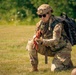A female military medic in full combat gear kneels on a grass field, appearing to attend to a casualty partially visible at the bottom right of the frame. She is wearing OCP (Operational Camouflage Pattern) uniform with a 'MED' patch visible on her shoulder, a tan tactical helmet, sunglasses, and carries a black MOLLE medical backpack. She is holding an orange training tourniquet, indicating this is likely a tactical combat casualty care (TCCC) training exercise.