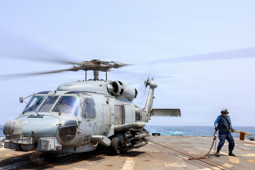 A military helicopter, identifiable as an MH-60R Seahawk, is on the flight deck of a naval vessel with its rotors spinning. A deck crew member in blue flight deck gear and helmet is handling a tie-down chain, likely preparing for or completing a landing/launch operation. The scene takes place at sea under overcast skies.