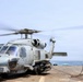 A military helicopter, identifiable as an MH-60R Seahawk, is on the flight deck of a naval vessel with its rotors spinning. A deck crew member in blue flight deck gear and helmet is handling a tie-down chain, likely preparing for or completing a landing/launch operation. The scene takes place at sea under overcast skies.