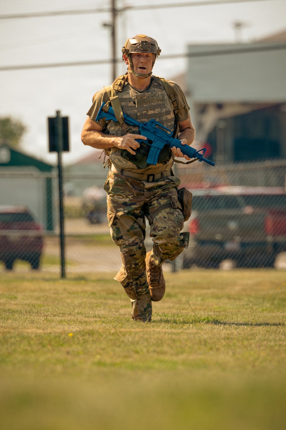 A military or law enforcement personnel in full tactical gear is running across a grassy field while carrying a bright blue training weapon. The individual is wearing OCP (Operational Camouflage Pattern) uniform, a tan tactical helmet, and a plate carrier vest. The blue-colored firearm indicates this is a training exercise using a simulated or inert weapon replica.